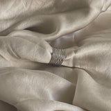 En sølvring samler lyse silkestofbølger i en blød, afslappet foldning. Stoffet skaber en glat og elegant tekstur i en neutral setting.