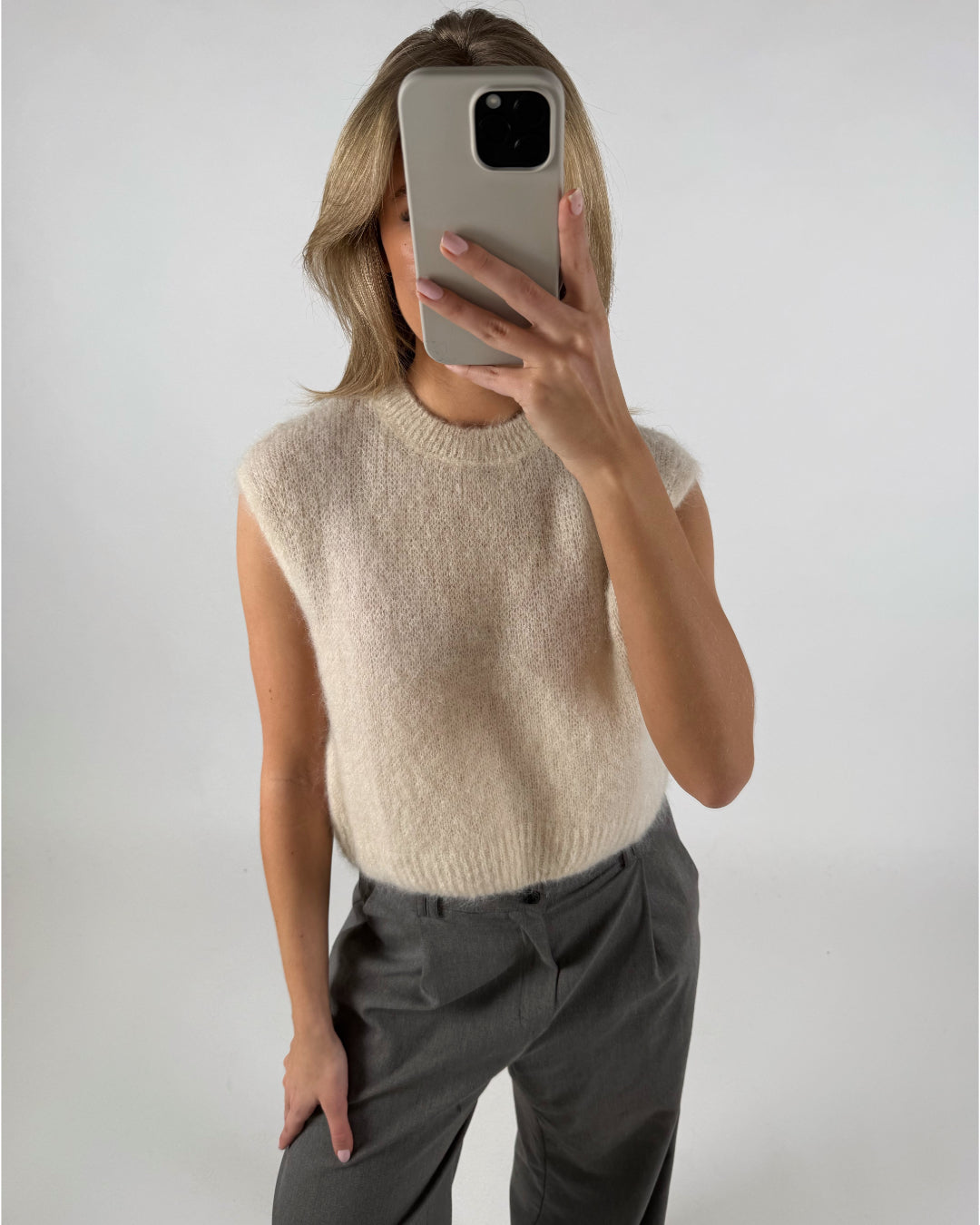 Person tager en selfie iført lyse ærmeløs sweater og grå bukser i et neutralt oplyst rum, dækkende ansigtet med en smartphone.