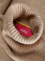 En lys brun rullekrave sweater folder sig, hvor en rød etiket med hvid tekst "JJXX" stikker ud fra kraven. En gul snor er også synlig.