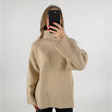 En kvinde tager et selfie med en sort mobiltelefon foran sit ansigt. Hun har en beige rullekrave sweater på og står foran en hvid baggrund.