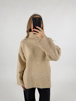 En kvinde tager et selfie med en sort mobiltelefon foran sit ansigt. Hun har en beige rullekrave sweater på og står foran en hvid baggrund.