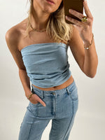 En person iført en stropløs blå top og matchende jeans tager et selfie i et spejl. Personen bærer guldsmykker og står i en lys baggrund.