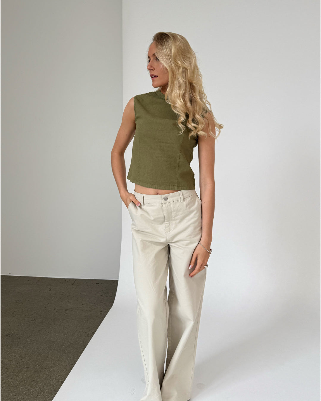 En kvinde med langt, blondt hår står iført en olivengrøn ærmeløs top og beige bukser i et minimalistisk indendørs rum med en hvid væg bagved.