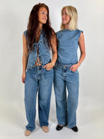To kvinder iført denim toppe og jeans står smilende og kigger på hinanden i et neutralt rum. Den ene har en blonde-top uden ærmer, den anden en ærmeløs bluse.