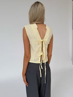 En kvinde med langt blond hår bærer en gul bluse med åbent rygdesign bundet med bånd, står stille i et minimalistisk rum med en rolig baggrund.