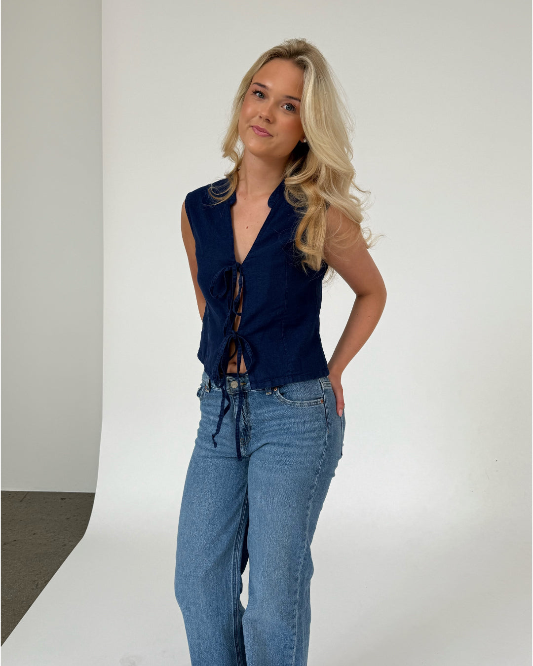 Kvinde står iført en sort, ærmeløs top og blå jeans i en studiolignende baggrund med en lys hvid væg og grå gulv. Hun ser roligt mod kameraet.