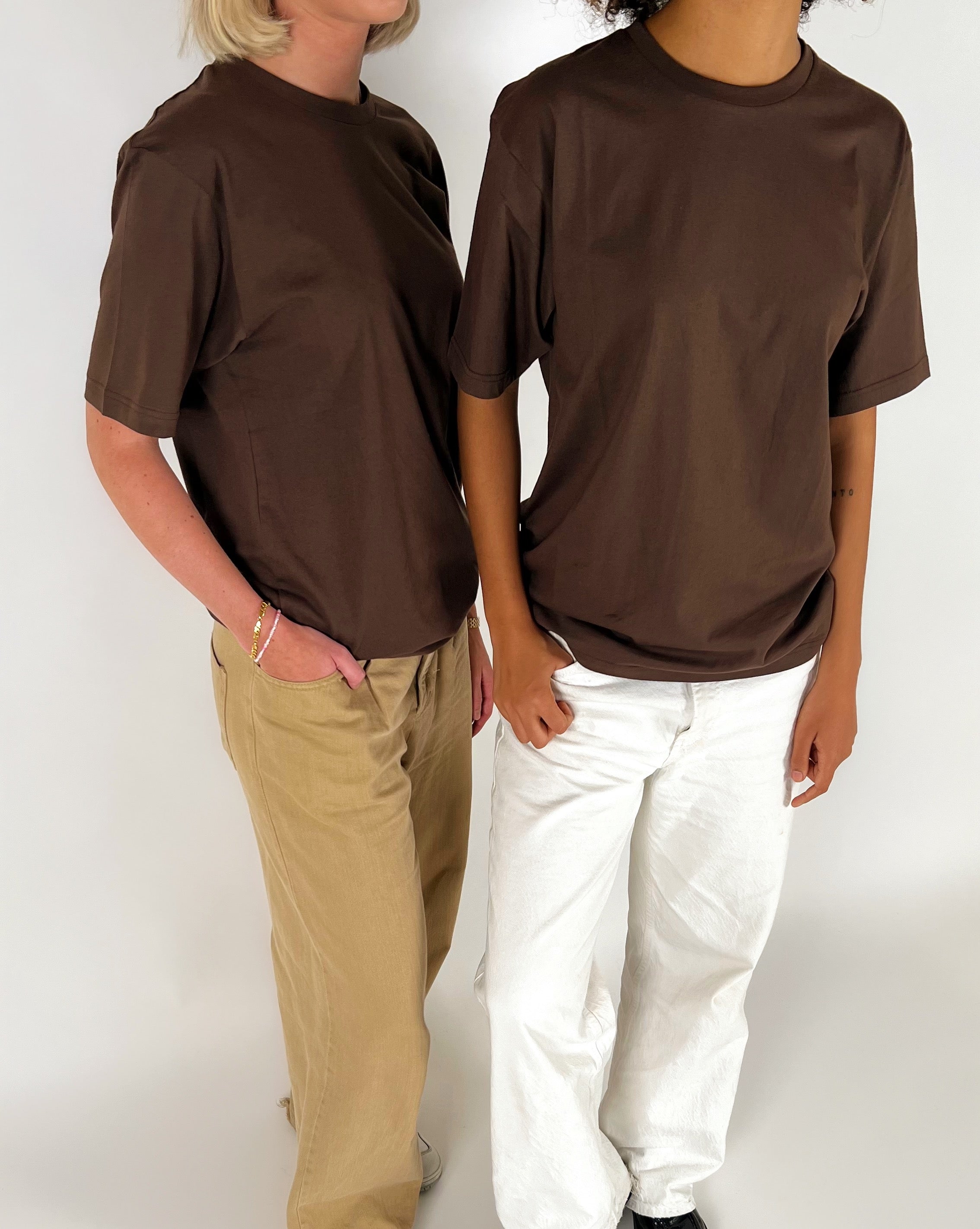 To personer iført brune t-shirts står i et neutralt rum. Den ene har beige bukser, den anden hvide. Begge står afslappet, delvist vendt mod hinanden.