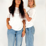 To kvinder i hvide T-shirts og blå jeans står sammen mod en hvid baggrund. De bærer smykker og udstråler selvsikkerhed.