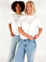 To personer i hvide T-shirts og jeans står smilende i et studie med hvid baggrund. Den ene person har krøllet hår, den anden har skulderlangt blond hår.