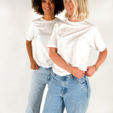 To kvinder iført hvide T-shirts og blå jeans står ved siden af hinanden og smiler i et lyst studiemiljø.