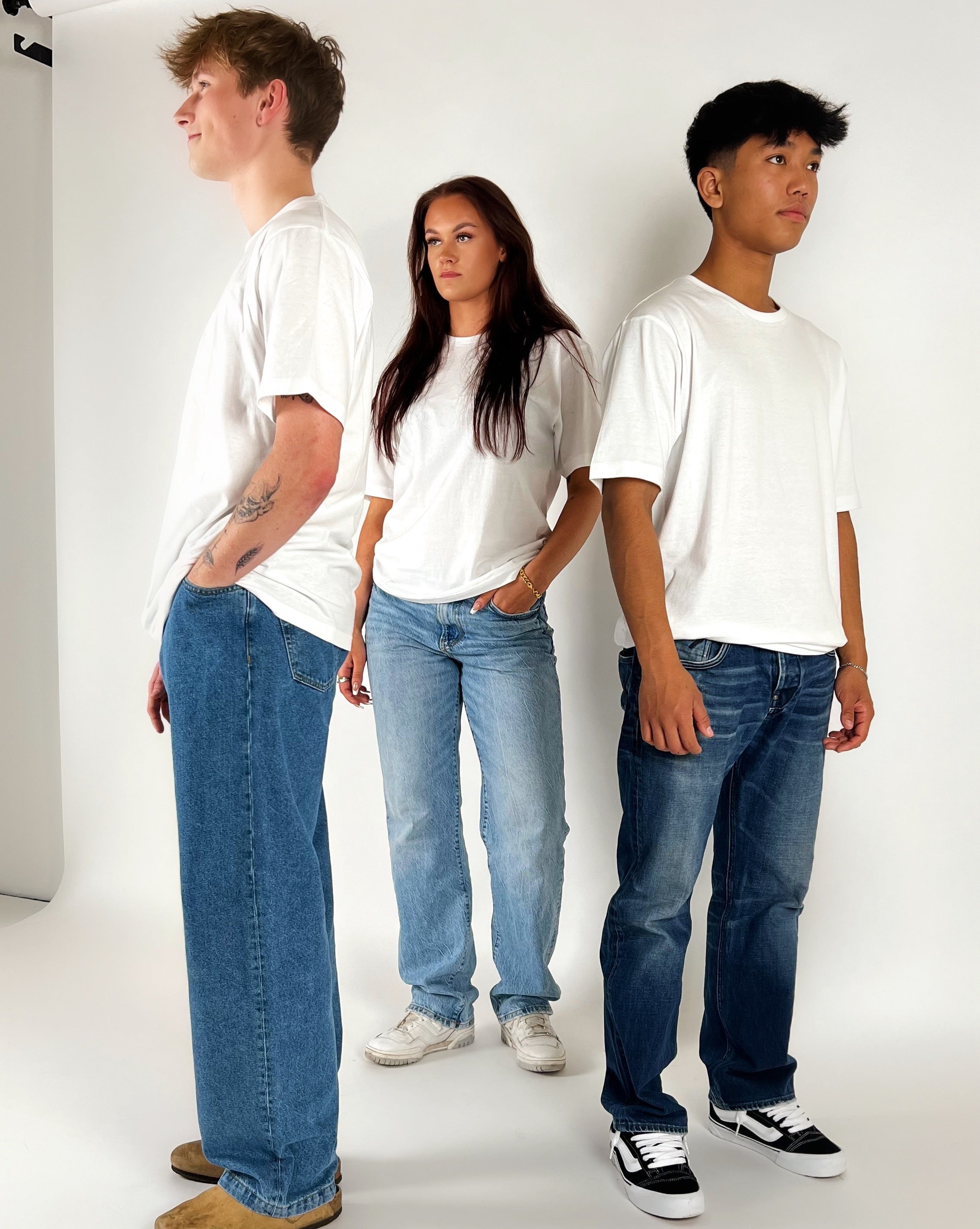 Tre personer står i studie iført hvide T-shirts og blå jeans, med neutral baggrund og lys belysning. To mænd står på hver sin side af kvinden.