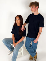 En person sidder på en hvid stol, iført en sort t-shirt og jeans, mens en anden person står ved siden af og hviler en arm på stolens ryglæn. Begge er i et lyst rum.