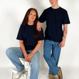 To personer i sorte t-shirts og blå jeans; den ene sidder på en hvid skammel, mens den anden står ved siden af i et lyst rum.