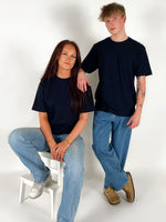 To personer i sorte t-shirts og blå jeans; den ene sidder på en hvid skammel, mens den anden står ved siden af i et lyst rum.
