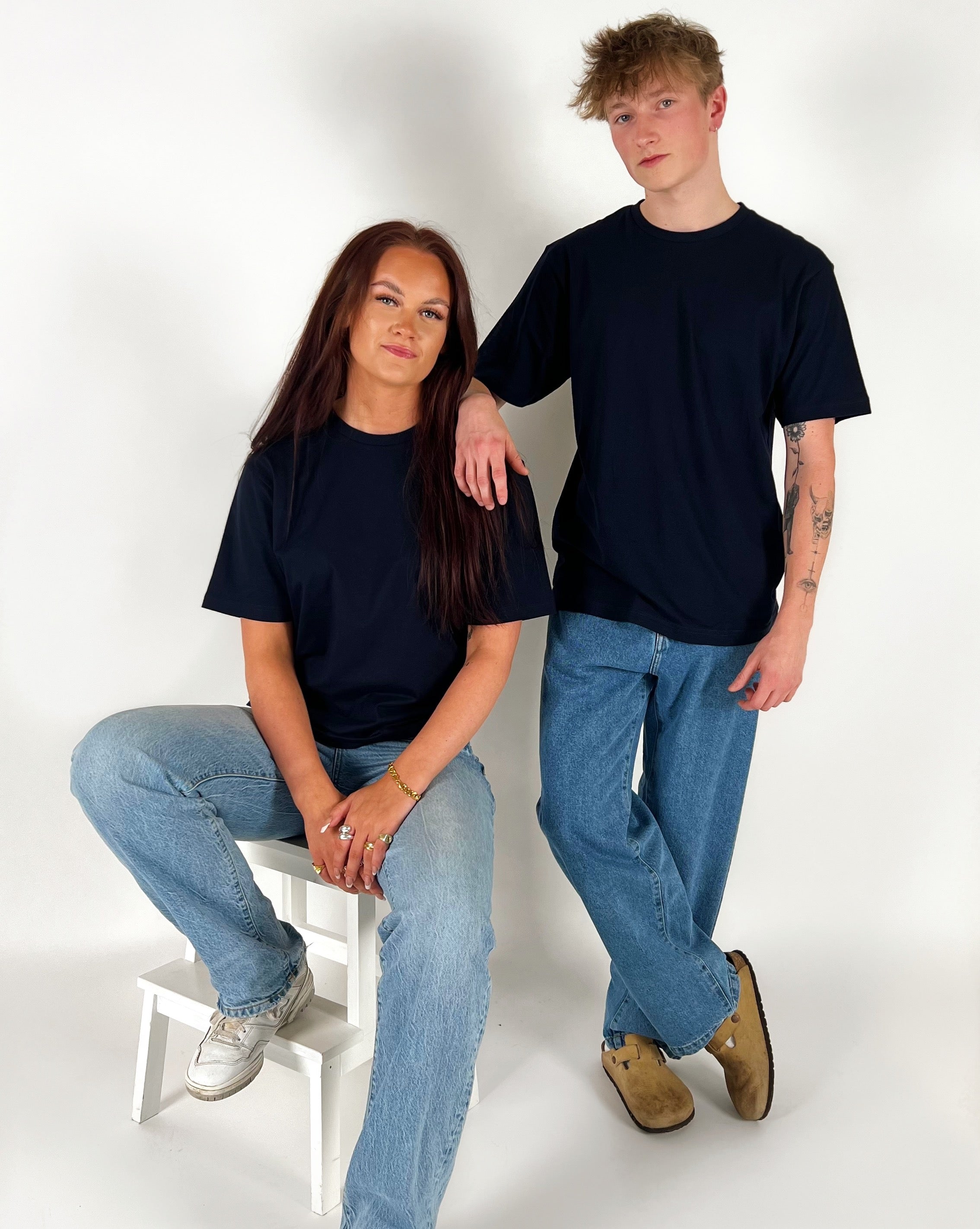 To personer i sorte t-shirts og blå jeans; den ene sidder på en hvid skammel, mens den anden står ved siden af i et lyst rum.