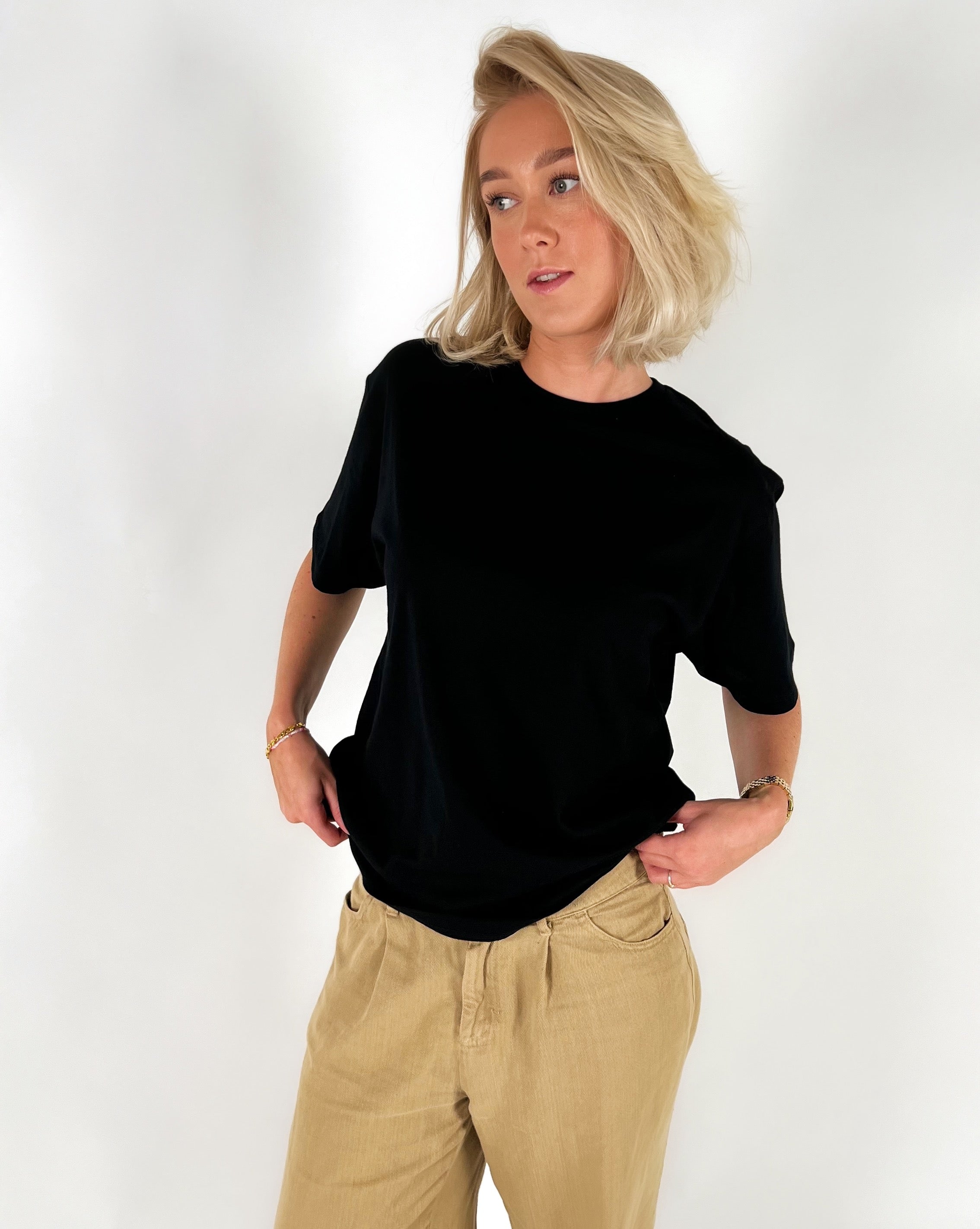 Person i sort t-shirt står med hænderne i lommerne, iført lyse bukser, med hvid baggrund.