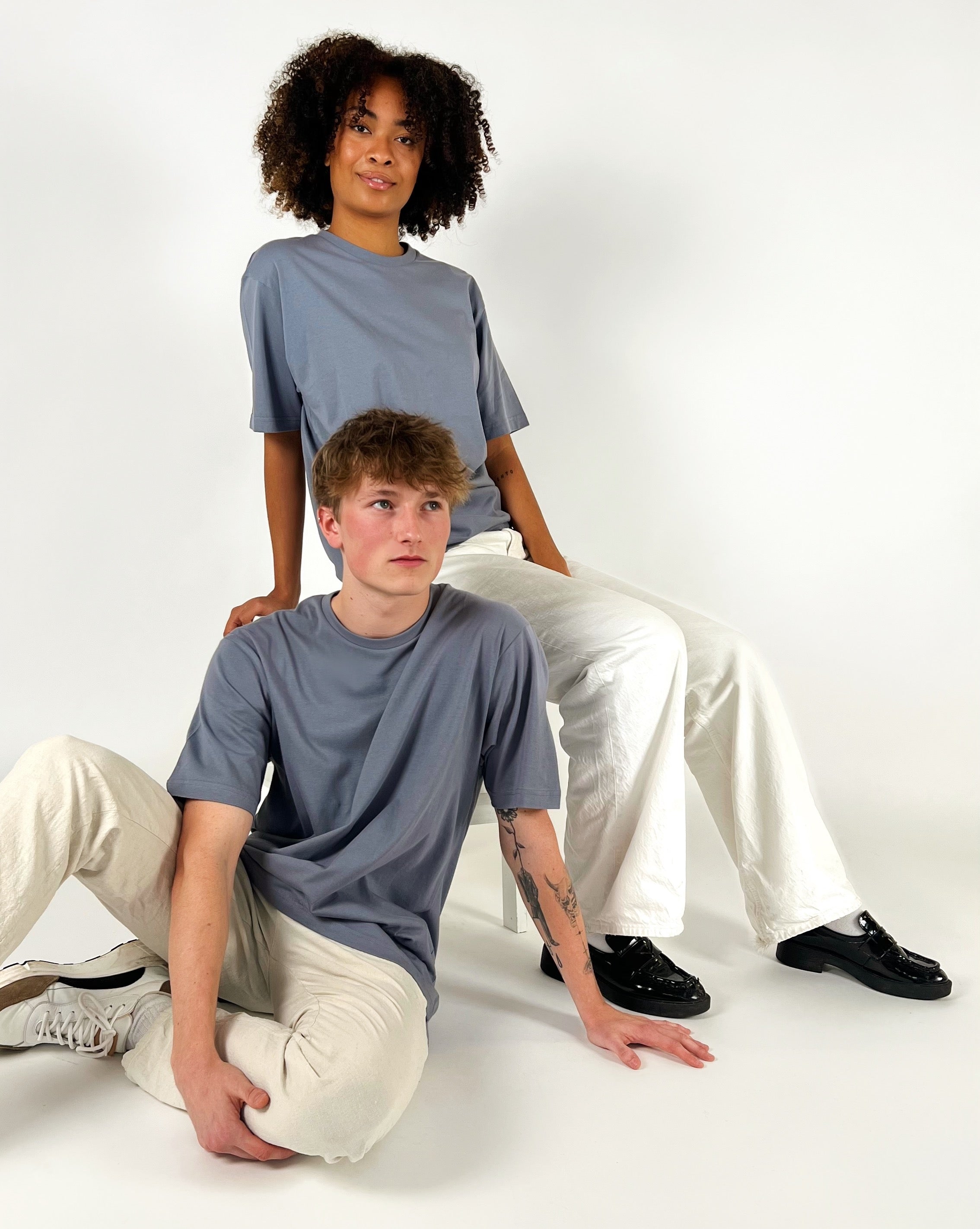 To personer i grå t-shirts og hvide bukser poserer i et studie. Den ene sidder på gulvet, den anden sidder på en stol. Baggrunden er hvid.