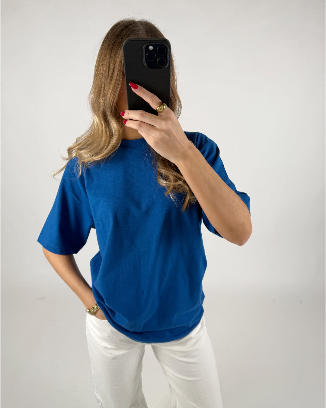En person iført en blå T-shirt tager et billede med en smartphone foran sit ansigt. Personen har langt hår og står i et neutralt rum.