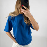 En person i en blå t-shirt holder en smartphone op foran ansigtet i en selfie-position. Personen står i et lyst rum med hvid baggrund.
