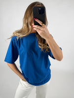 En person i en blå t-shirt holder en smartphone op foran ansigtet i en selfie-position. Personen står i et lyst rum med hvid baggrund.