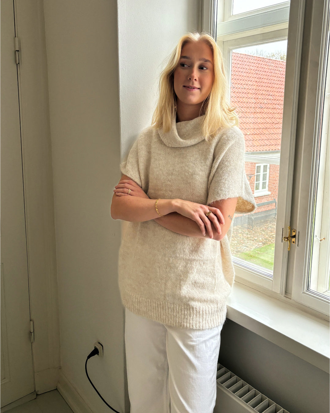 En person står med krydsede arme iført en lys sweater og hvide bukser. De står ved et vindue med udsigt til et rødt hustag og hvidmalede vægge.