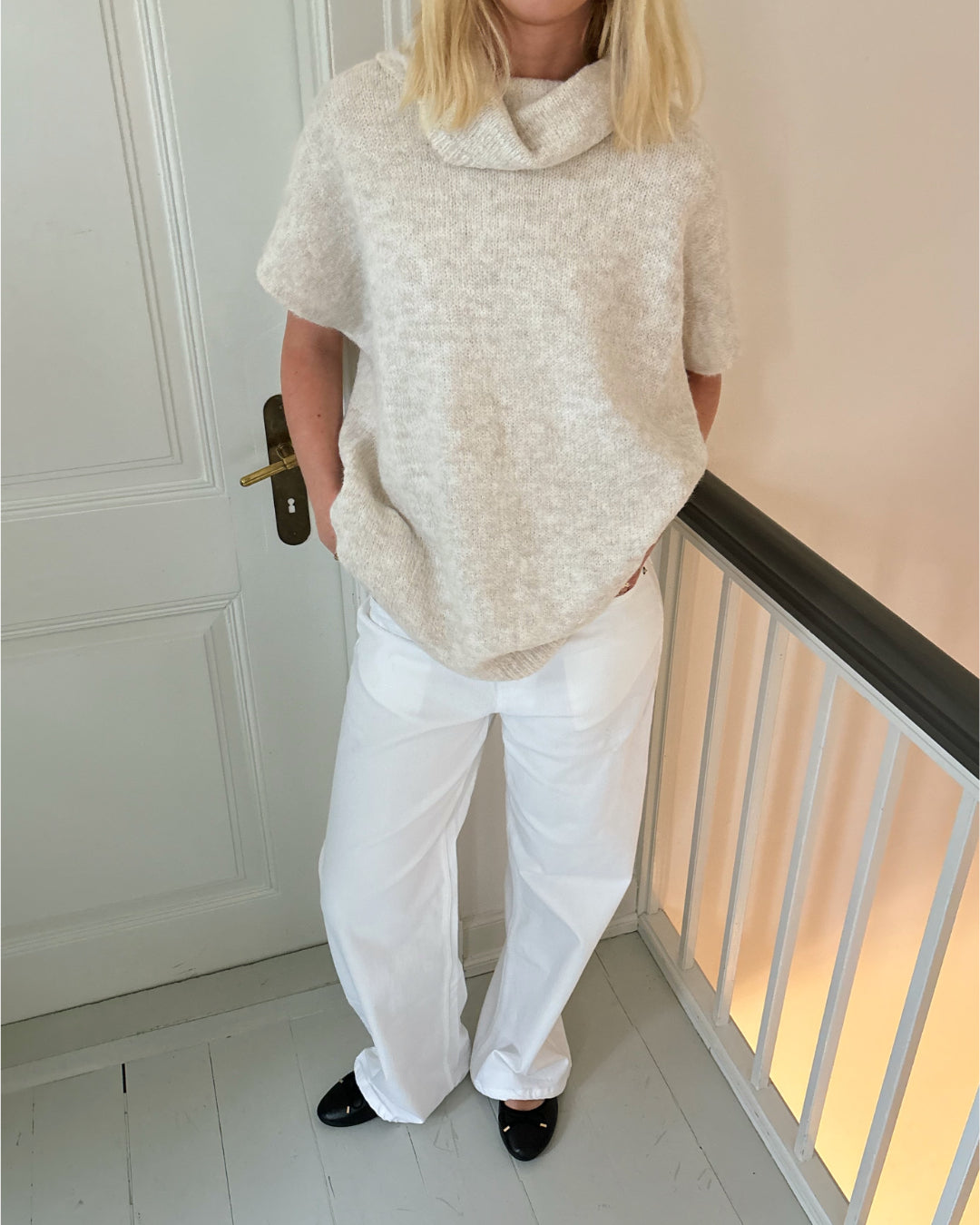En person iført en beige sweater og hvide bukser står med hænderne i lommerne ved siden af en hvid dør i et lyst rum med gelænder.