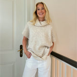 En person står og smiler iført en beige sweater og hvide bukser. Personen læner sig mod en lys væg ved siden af en dør.
