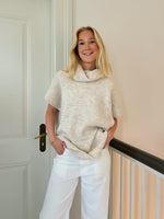 En person står og smiler iført en beige sweater og hvide bukser. Personen læner sig mod en lys væg ved siden af en dør.