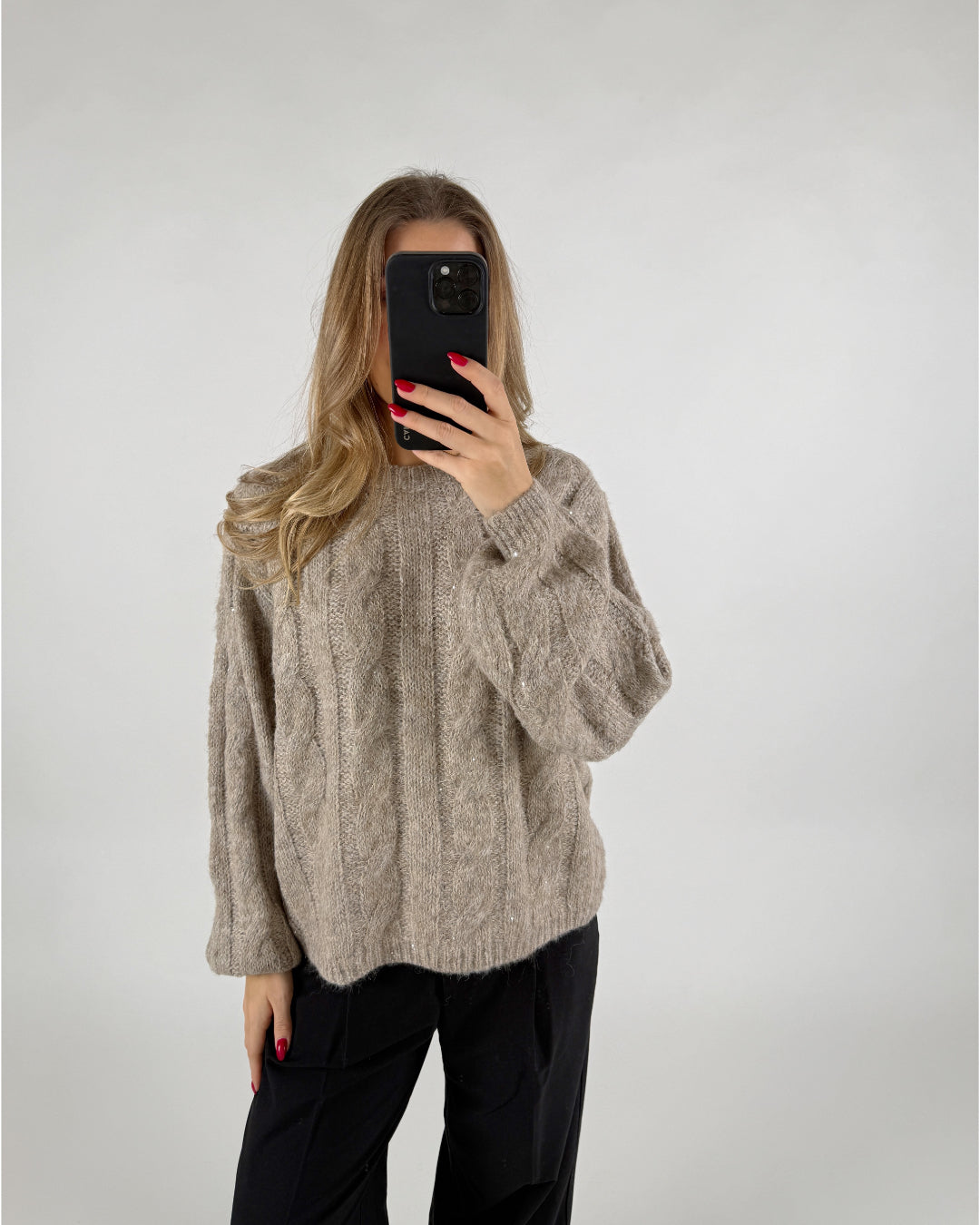 En person iført en beige striktrøje tager et billede af sig selv med en smartphone. Personen står i et rum med en neutral, lys baggrund.