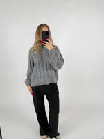 Person tager et spejlbillede iført en grå striktrøje og sorte bukser. Personen holder en telefon med rødnegler i et minimalistisk, lyst rum.