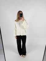 En person iført en hvid sweater og sorte bukser tager et selfie foran et spejl i et minimalistisk rum med hvid baggrund.