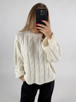 Kvinde iført hvid sweater tager selfie med telefon foran ansigt; langærmet trøje med flettemønster. Hendes negle er malet rød, og hun står mod en neutral baggrund.