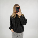 En person tager et billede med en smartphone i et spejlløst rum. De bærer en sort sweater og grå bukser, med hånden i lommen. Baggrunden er enkel og lys.