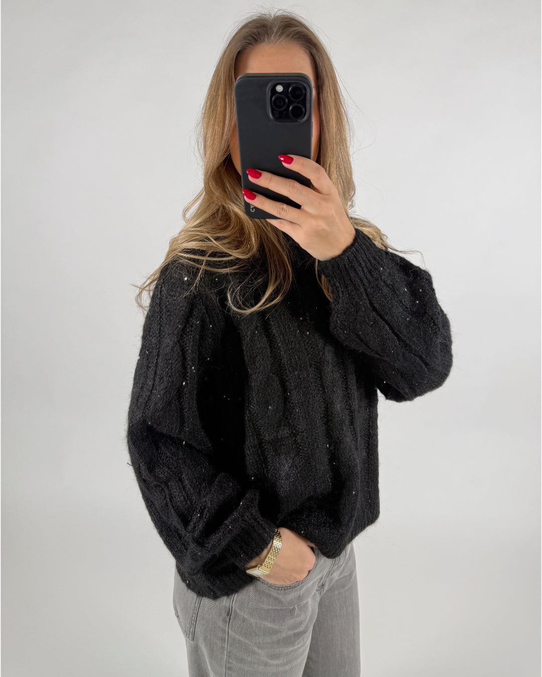En person iført en sort striktrøje tager et billede med en sort smartphone. De har løst hår og røde negle. Baggrunden er enkel og lys. Ingen tekst til stede.