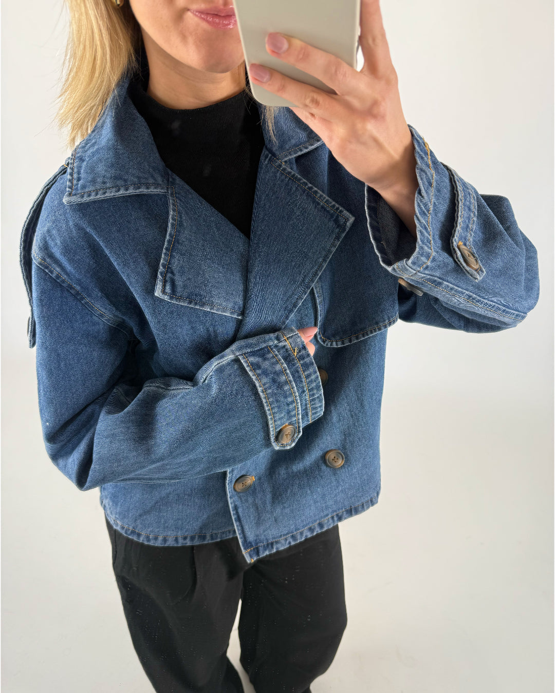 En person iført en blå denimjakke tager et spejlbillede med en smartphone. Personen har lyst hår og bærer en sort trøje under jakken i et neutralt rum.