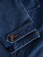 En blå denimjakke har en knap fastgjort på ærmet. Stoffet er detaljeret med orange syninger, der skaber et klassisk og robust look.