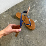En hånd holder en brun sandal med blå stropper, mens den ligger på en grå betonoverflade ved siden af en hvid væg.