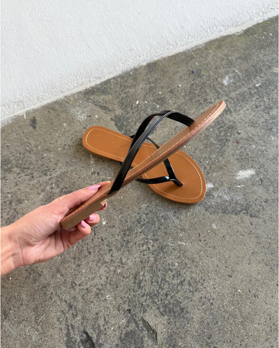 En brun og sort flip-flop sandal holdes i hånden over en grov betonoverflade, mens en anden sandal ligger på gulvet ved siden af en hvid væg.