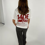 En person står med ryggen til iført en hvid T-shirt med rød grafik af en bil og tekst "140E Racing Club member 140E ... 1987 ... Car racing ...". De er i en lys indendørs omgivelser.