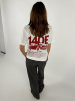 En person står med ryggen til iført en hvid T-shirt med rød grafik af en bil og tekst "140E Racing Club member 140E ... 1987 ... Car racing ...". De er i en lys indendørs omgivelser.