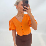 En person tager en selfie iført en orange bluse og brun nederdel, stående foran en lys baggrund. Personen holder en mobiltelefon, og bærer smykker.