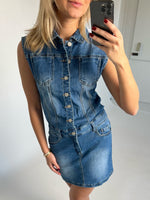 Kvinde i en ærmeløs denimkjole tager et spejlfoto af sig selv. Hun har rødmalede negle og et armbånd. Baggrunden er et lyst rum med simple vægge.