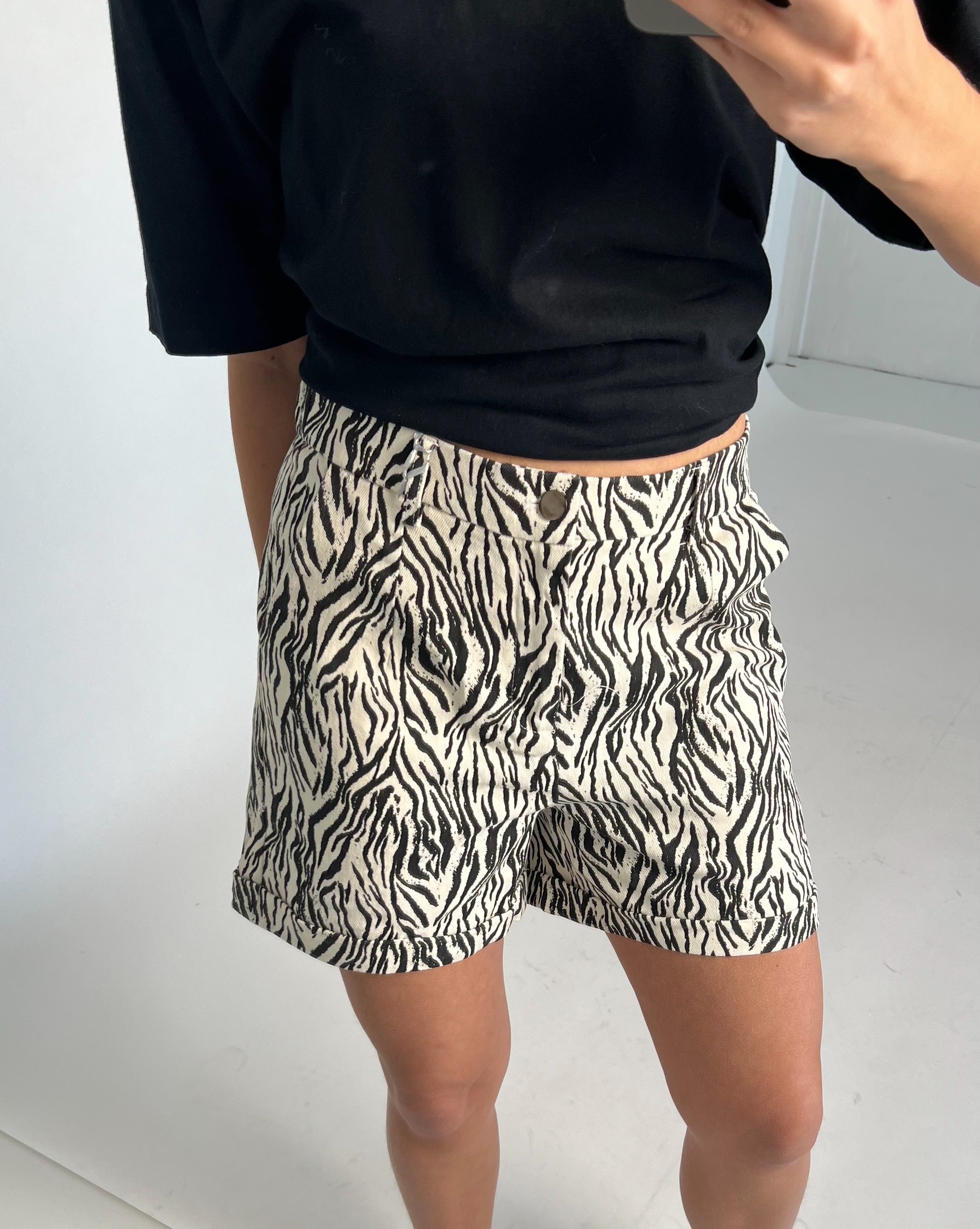 En person iført en sort trøje fotograferer sig selv iført zebrastribede shorts i et minimalistisk, hvidt rum.
