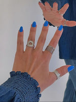 Hånd med blå neglelak strækker sig mod et spejl, iført flere ringe og en bluse med blå ærmer; reflekteres i en spejlflade.