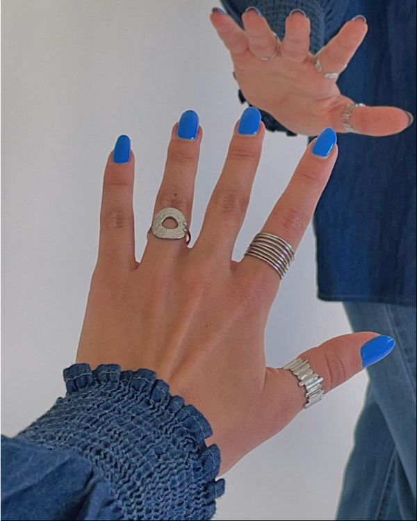 Hånd med blå neglelak strækker sig mod et spejl, iført flere ringe og en bluse med blå ærmer; reflekteres i en spejlflade.
