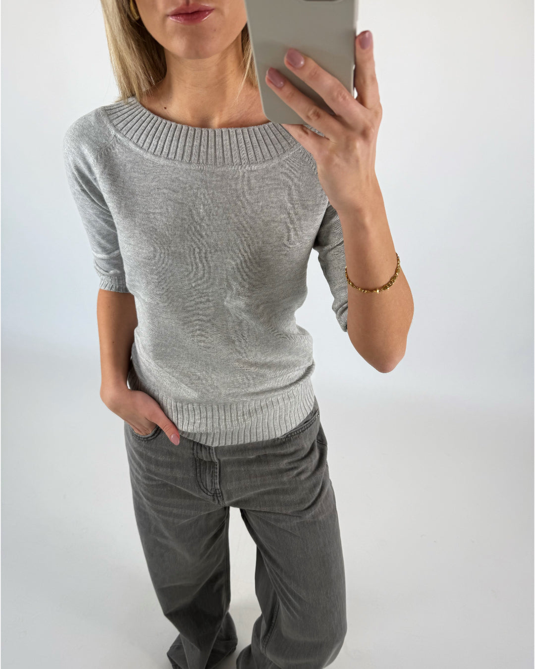 Kvinde iført grå sweater tager et selfie i spejl. Hun har hånden i lommen på sine grå bukser. Baggrunden er ensfarvet lys.