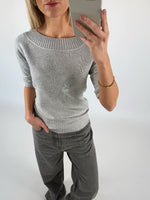 Kvinde iført grå sweater tager et selfie i spejl. Hun har hånden i lommen på sine grå bukser. Baggrunden er ensfarvet lys.