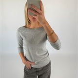 En person tager en selfie foran en hvid baggrund, iført en grå sweater og jeans, med hånden i lommen og en mobiltelefon foran ansigtet.