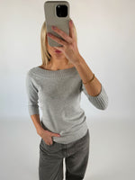 En person tager en selfie foran en hvid baggrund, iført en grå sweater og jeans, med hånden i lommen og en mobiltelefon foran ansigtet.