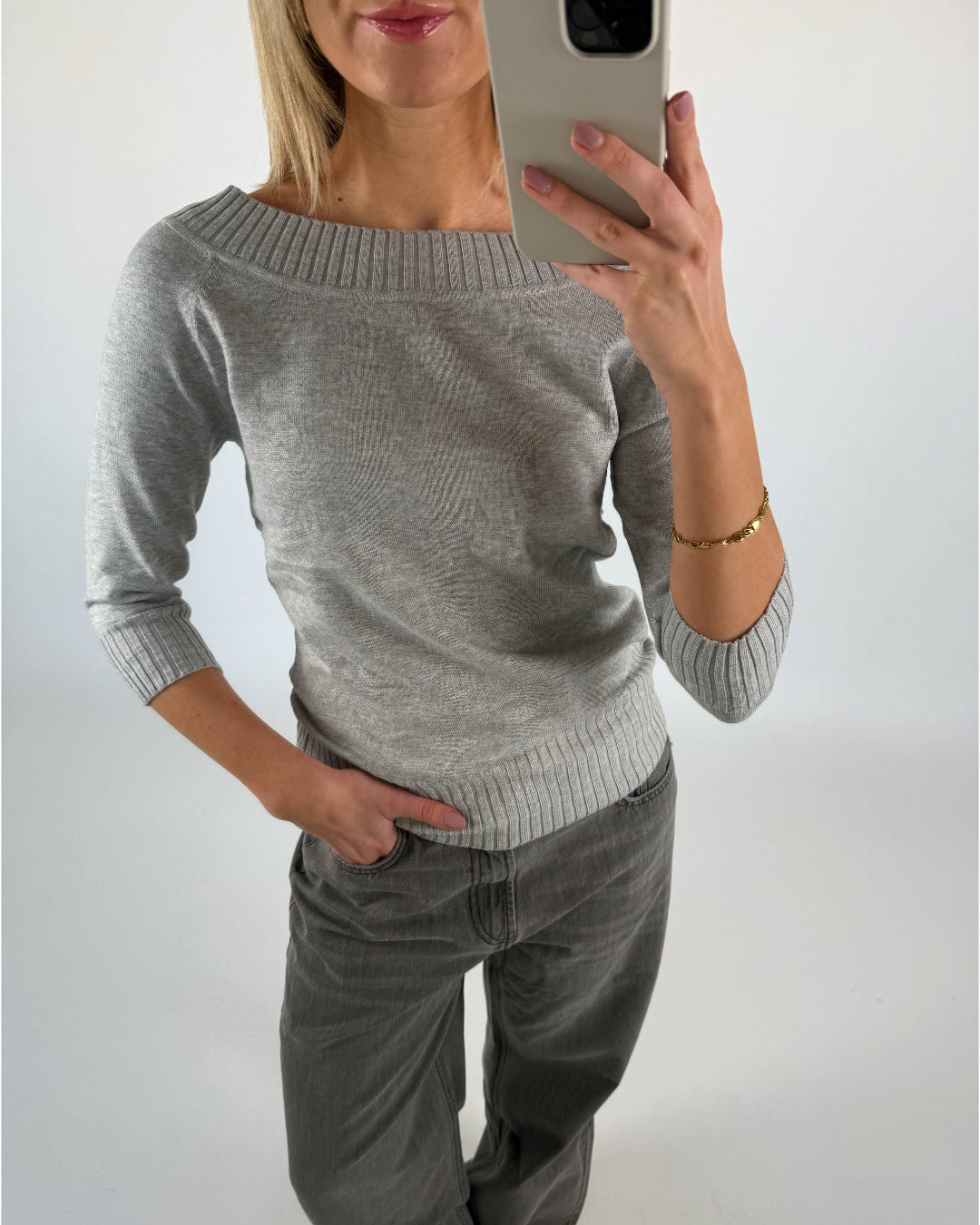 En person tager en selfie i et spejl. De bærer en grå sweater og bukser, står med én hånd i lommen og har et guldarmbånd. Baggrunden er neutral.